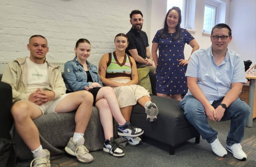 A group of 6 young people and staff members sat on a sofa smiling at camera. 3 female and 3 male.