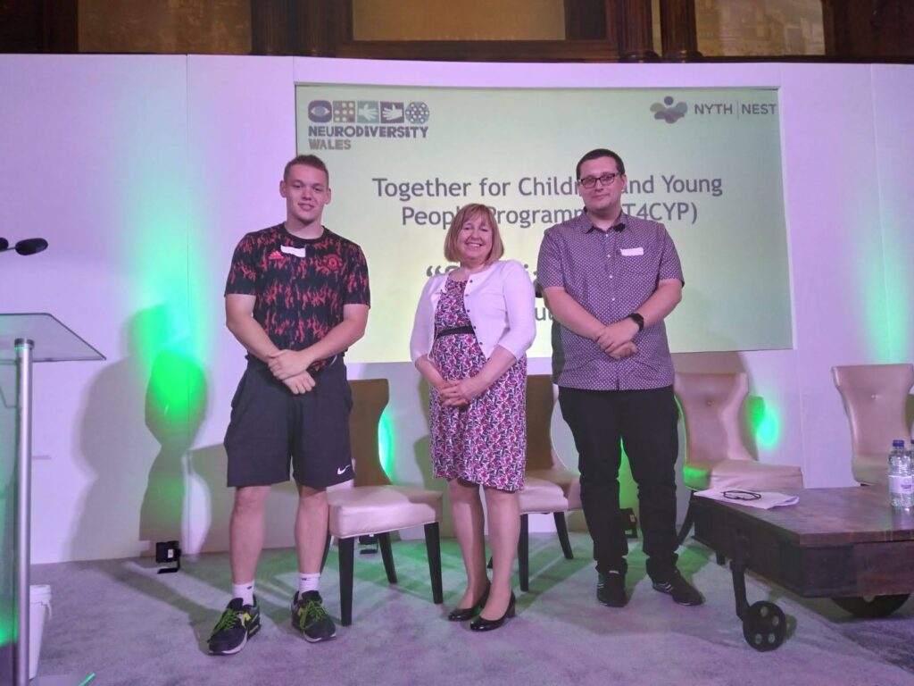 Minister Lynne Neagle with two young men standing each side of her. A power point presentation is behind them