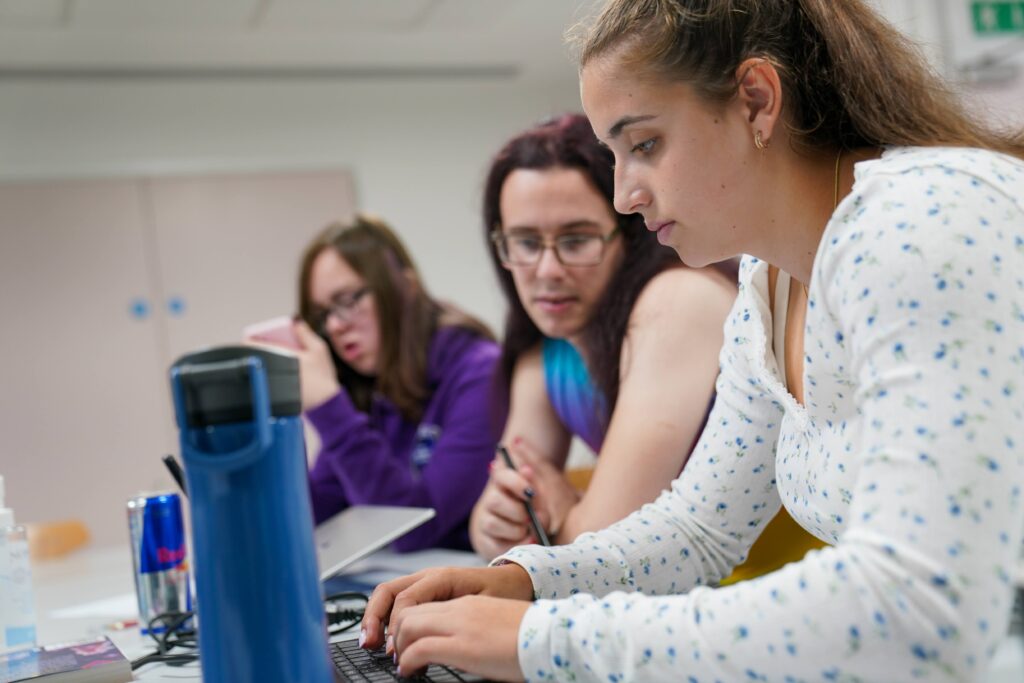 Three young people sat at a table working together. One us using the laptop. 