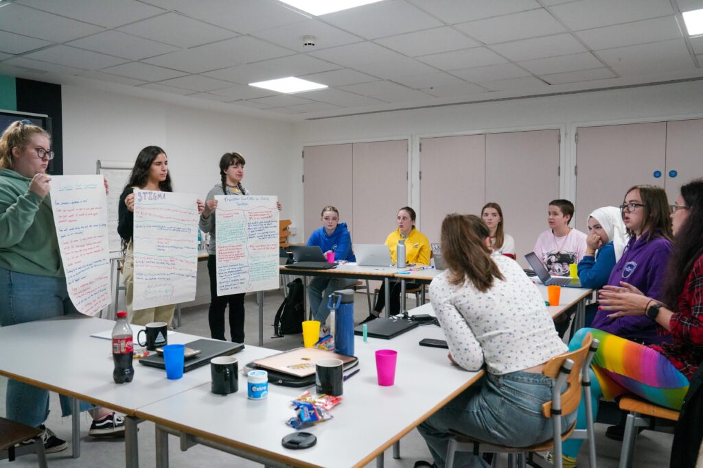 Three staff members stood presenting on flip chart paper to a room of young people. 