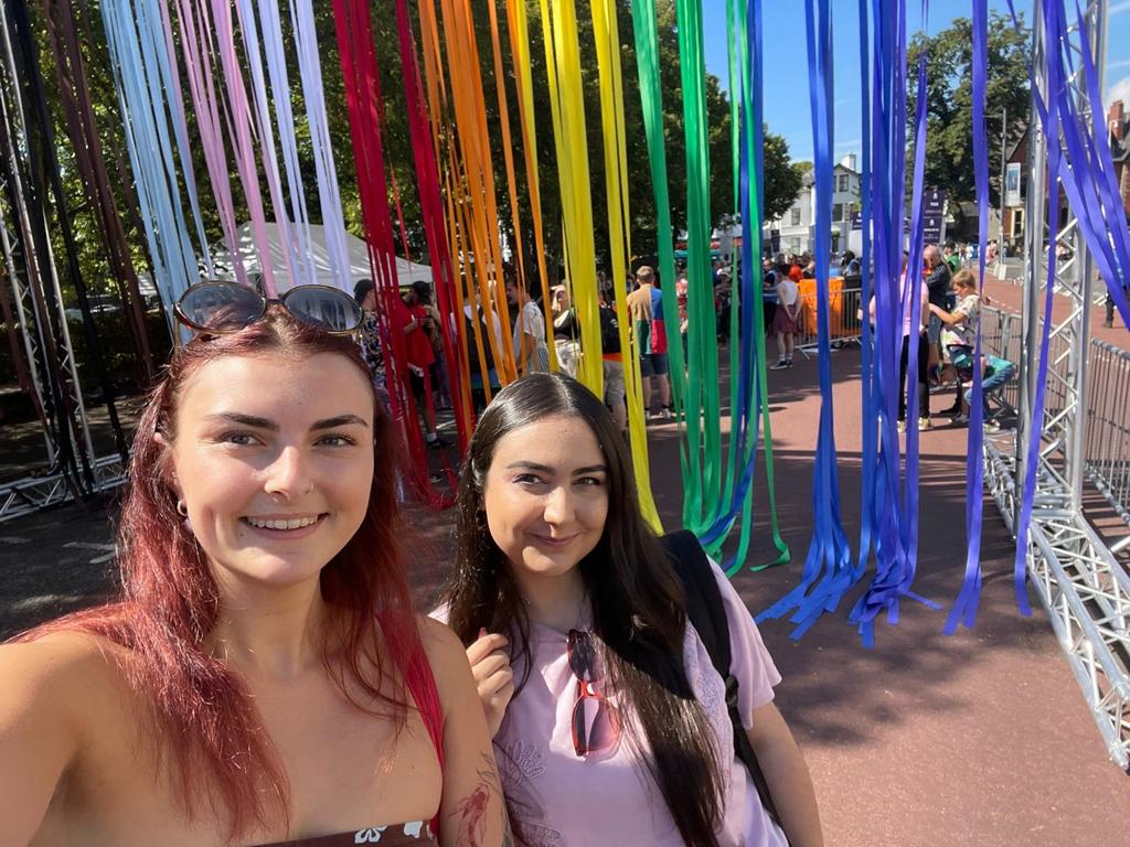 Two women talking a selfie in front of a rainbow background made of ribbons