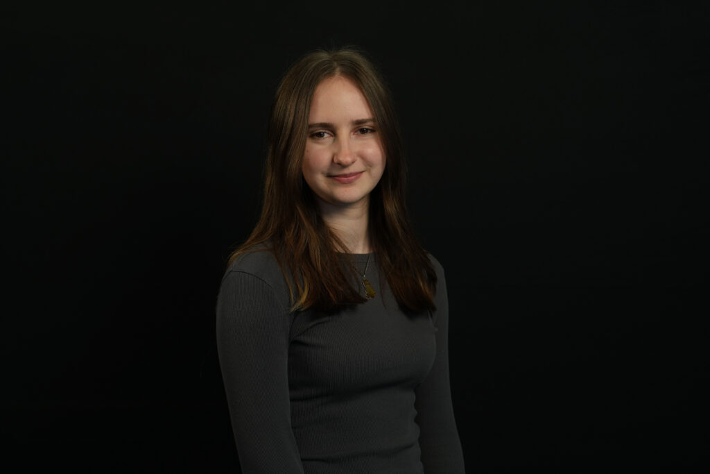 A young female with brown hair and a charcoal grey long sleeve top stands infront of a black background. 