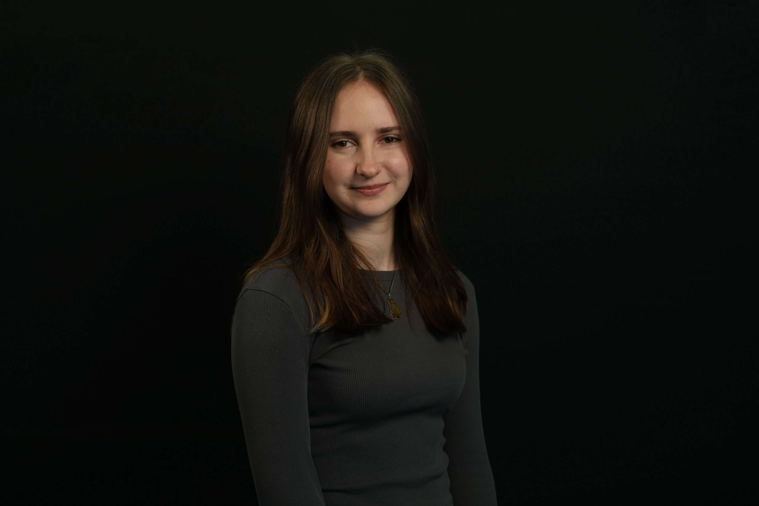 A young female with brown hair and a charcoal grey long sleeve top stands infront of a black background.